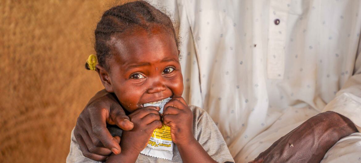 Un jeune enfant africain nommé Naba est montré en train de consommer des compléments alimentaires nutritionnels à Adré, Ouaddai, Tchad. Elle est tenue par les mains d'un adulte et sourit tout en mangeant le sachet de nourriture.