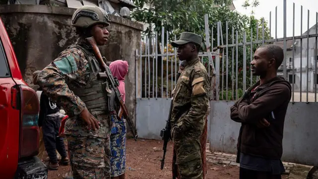 Deux soldats en tenue militaire discutant avec un civil, femmes en arrière-plan